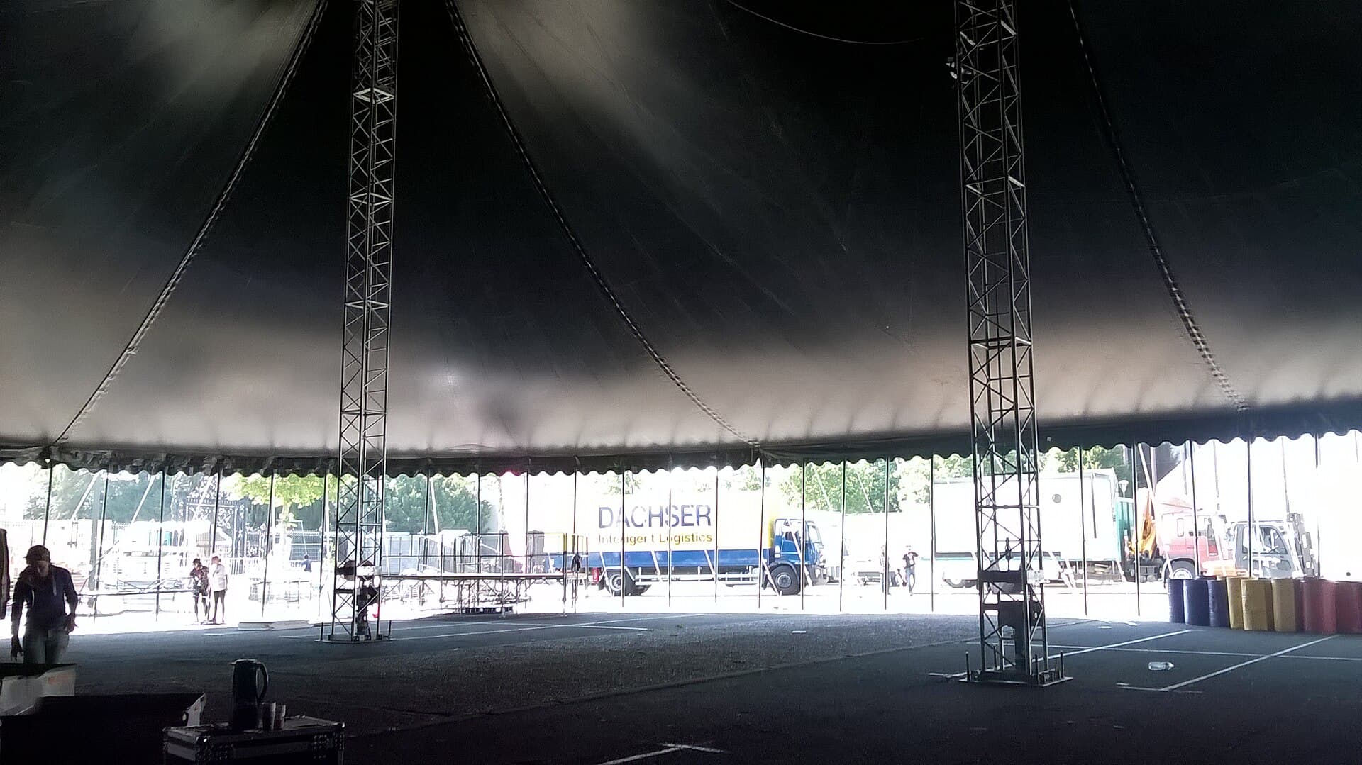 L'intérieur d'un grand chapiteau en cours de montage : mâts, structures métalliques et camions de livraison en arrière-plan.