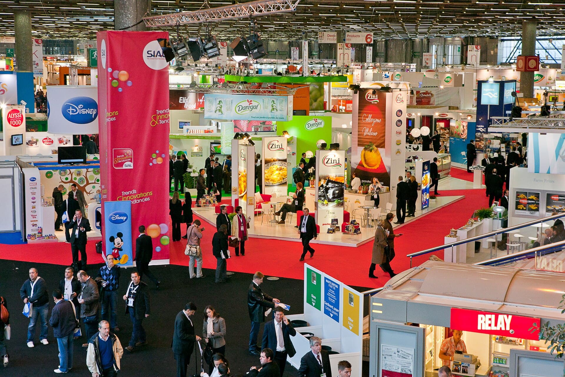 Un hall de salon professionnel vu d'en haut : allées à moquette rouge, stands et visiteurs.